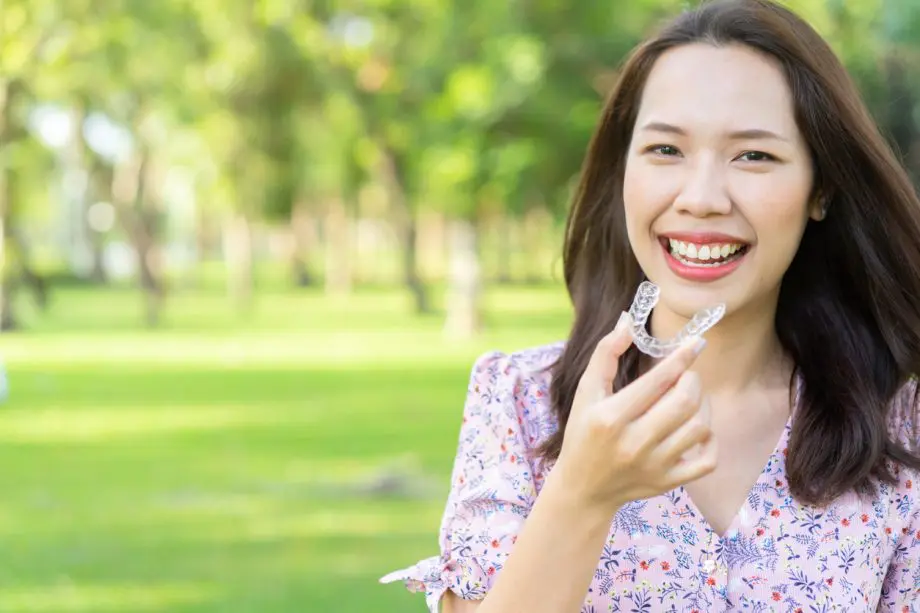 Woman with straight brown hair holding a pair of Invisalign aligners while sitting in a park on a sunny day.