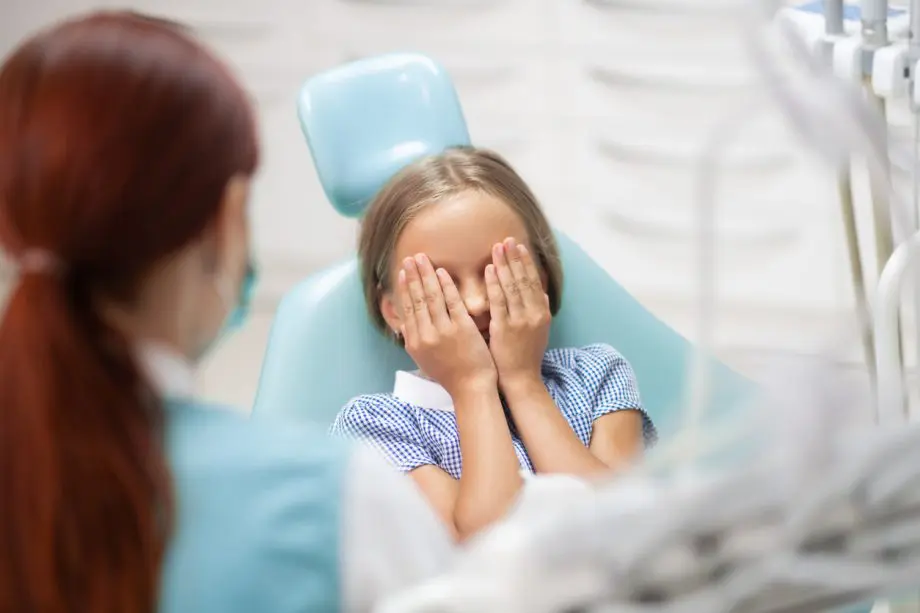 Photograph of a young girl sitting in a dentists chair with her eyes covered as a female hygienist is nearby.