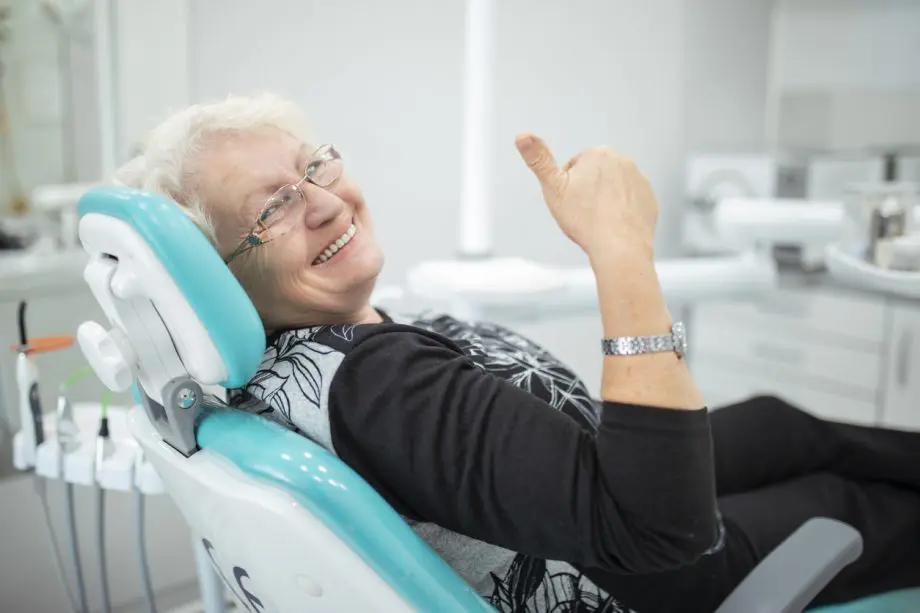 Photograph of an older woman with short white hair sitting in a dentists chair smiling and giving a thumbs-up.