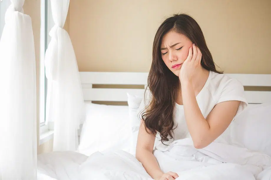 woman holds jaw and grimaces in pain while sitting in bed