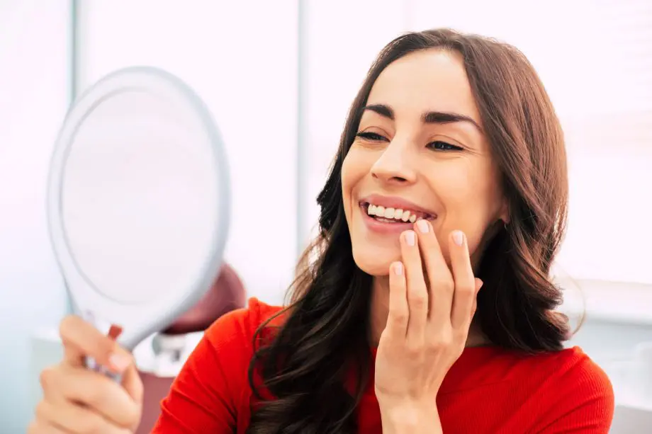 woman using hand held mirror to look at her teeth while smiling