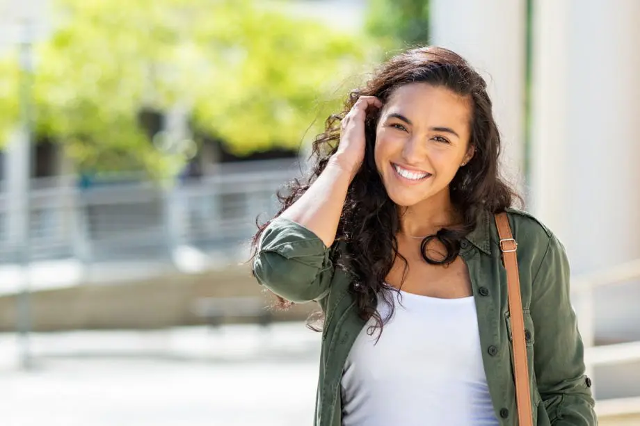 woman standing on sidewalk smiling, pushing her long hair behind her ear