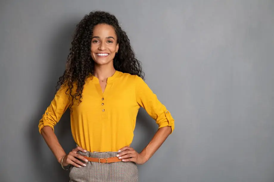 smiling woman in bright yellow shirt with hands on hips standing in front of grey backdrop